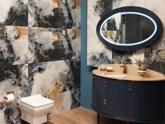 Mavi onyx görünümlü porselen duvarın önünde lacivert yivli banyo dolabı ve ışıklı oval ayna