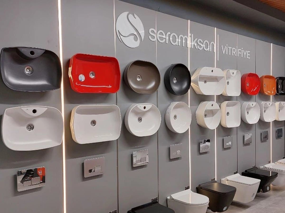 Seramiksan vitrifiye teşhir duvarı: iki sıra hâlinde renkli lavabolar, altlarında gömme rezervuar kapakları ve asma klozetler